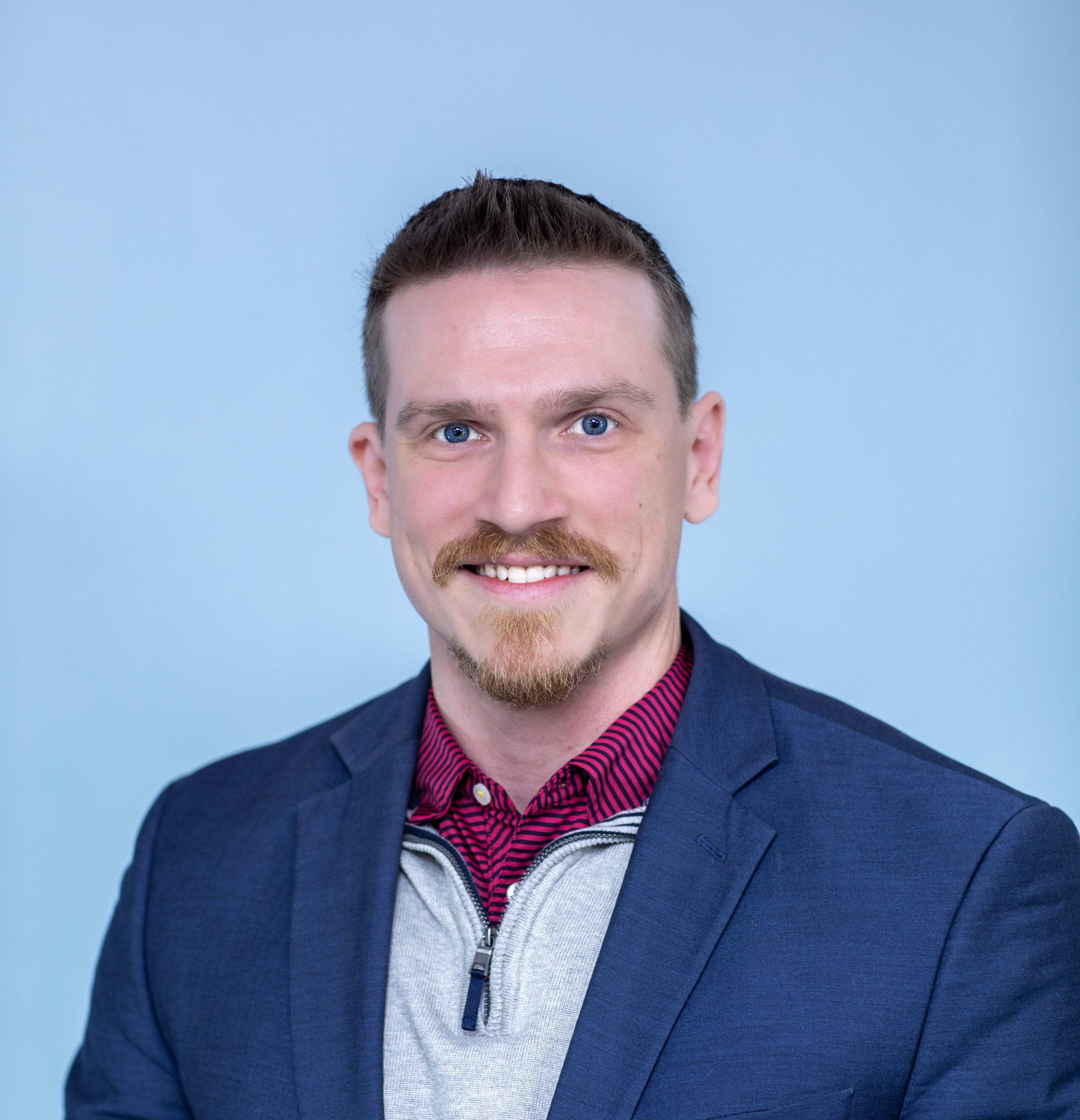 A man with short brown hair, a mustache, and a goatee, wearing a blue blazer, gray sweater, and red shirt, poses against a light blue background.